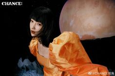 a woman in an orange dress standing next to a giant object with the moon behind her