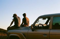 two men are sitting on the hood of a car looking at something in the distance