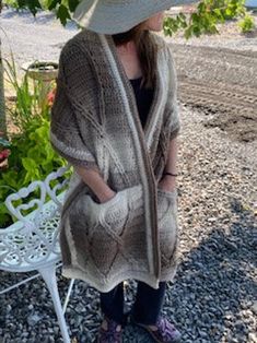 a woman wearing a hat and cardigan is standing on the gravel near a bench