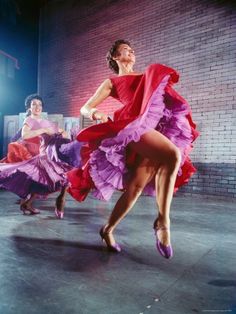 three women in colorful dresses are dancing on the stage with their legs spread out and one woman is wearing a red dress