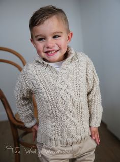 a young boy wearing a sweater and pants standing in front of a chair smiling at the camera