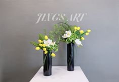 two black vases with yellow and white flowers in them on a table next to a gray wall