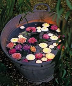 candles floating in a bucket filled with water surrounded by pink and red flowers on the ground