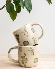 two coffee mugs with cactus designs on them sitting next to a potted plant