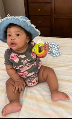 a baby sitting on a bed wearing a hat and holding a yellow toy in it's hand