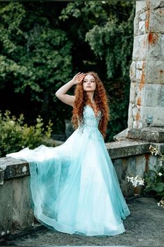 a woman with long red hair wearing a blue dress standing on a stone wall in front of trees