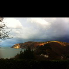 a view of the ocean and mountains from an overlook point on a cloudy day with sun coming through the clouds