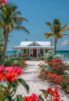 a white house on the beach surrounded by palm trees and red flowers in front of it