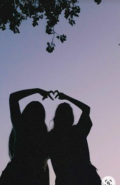 two women standing in front of a tree making a heart shape with their hands while the sun is setting