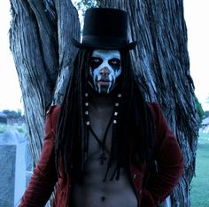 a man with white face paint wearing a top hat and long dreadlocks standing in front of a tree