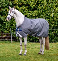 a white horse wearing a grey blanket standing in the middle of a grassy field with trees and bushes behind it