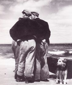 two men and a dog standing on the beach looking out at the ocean while another man stands next to them