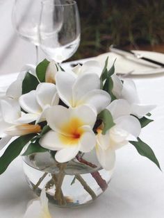 white flowers in a glass vase on a table with wine glasses and napkins behind it