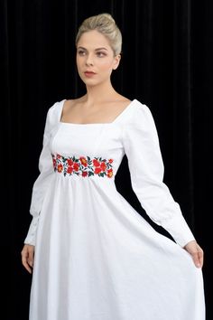 a woman wearing a white dress with red flowers on the chest and sleeves, standing in front of a black curtain