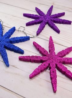 three purple and blue glittered star ornaments on a white wooden table with silver hooks