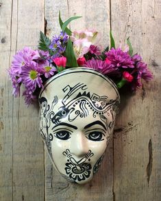 a white mask with flowers in it on a wooden wall next to purple and pink flowers