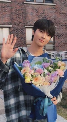 a young man holding flowers in front of a brick building with his hand up to the camera