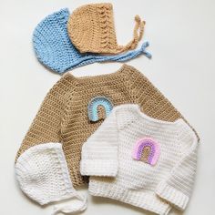 three crocheted sweaters and hats are shown on a white surface, including one with a rainbow in the middle