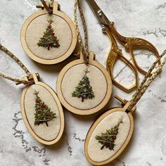 three cross stitch christmas tree ornaments hanging from gold chains on a marble surface with scissors