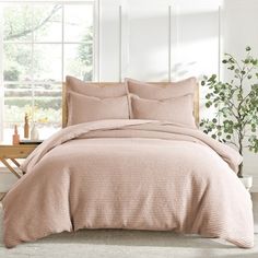 a bed with pink comforter and pillows in front of a large window next to a potted plant