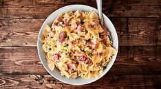 a white bowl filled with pasta and ham on top of a wooden table next to a fork