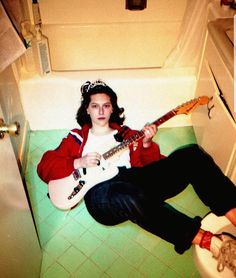 a person sitting on the floor with a guitar in their hand and a toilet behind them