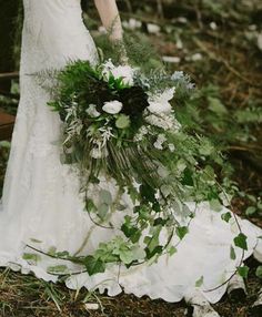 a woman standing in the woods holding a bouquet of greenery and white flowers on her wedding day