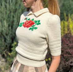 a woman with red hair wearing a white sweater and plaid skirt standing in front of bushes