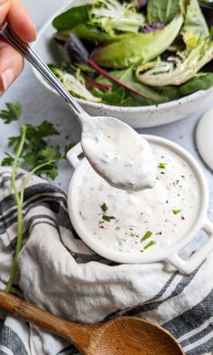 a hand holding a spoon full of ranch dressing next to a bowl of salad and a wooden spoon