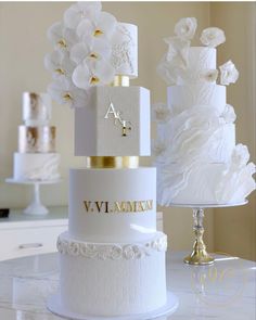 three tiered wedding cake with white flowers and gold lettering on the top, sitting on a table