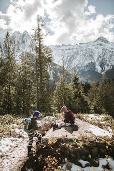 two people sitting on top of a rock in the middle of snow covered ground with mountains in the background