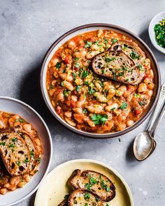 two bowls filled with beans and meat on top of a gray table next to silver spoons