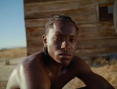 a man with dreadlocks sits in front of a wooden building and looks at the camera