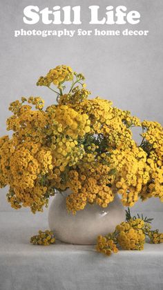 a white vase filled with yellow flowers sitting on top of a gray table next to the words still life photography for home decor