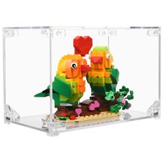 two lego birds in a glass case with plants and flowers inside it, one is orange and the other is green