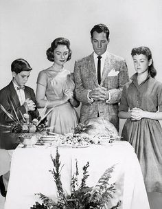 an old black and white photo of four people standing around a table with food on it