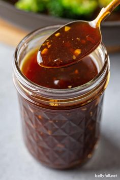 a spoon full of bbq sauce on top of a plate with broccoli in the background