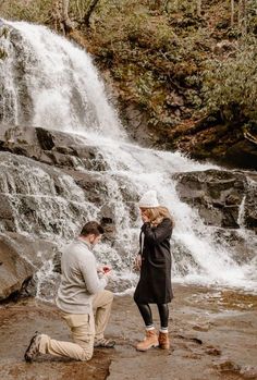 a man kneeling down next to a woman in front of a waterfall with a phone