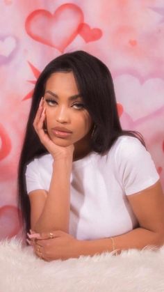 a woman sitting in front of a pink wall with hearts on it and her hand under her chin