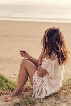 a woman sitting on top of a sandy beach next to the ocean holding a cell phone