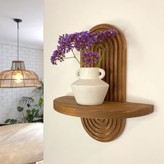 a white vase with purple flowers sitting on a wooden shelf next to a light fixture