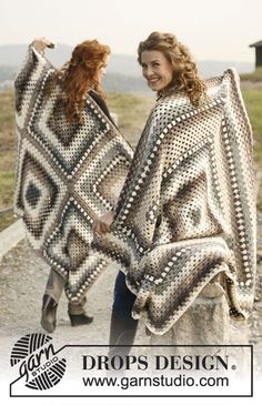 two women walking down a dirt road holding up some knitted shawls in front of them