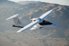 a small white airplane flying through the air over a field and mountain range in the background