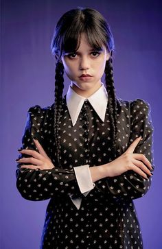 a woman with her arms crossed wearing a black and white polka dot dress, standing in front of a purple background