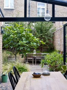 a wooden table sitting under a glass roof next to a lush green tree filled garden