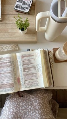 an open book sitting on top of a wooden table next to a cup and plant