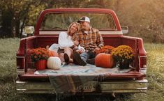 a man and woman sitting on the back of a red pickup truck with pumpkins