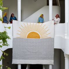 three women standing on the balcony of a building with a large quilt hanging from it's side