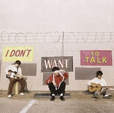 three men sitting on the side of a road with guitars in front of barbed wire