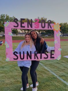 two girls holding up a pink sign that says sunrise on the side of a field